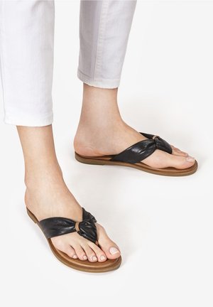 Pieds portant des sandales en cuir noir à nœud sur le dessus avec semelles plates marron, associées à un pantalon blanc court sur fond blanc.