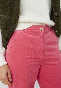 Pantalons en velours côtelé rose avec une texture à côtes verticales et une fermeture à bouton, associés à un pull en tricot blanc et un gilet vert olive.