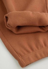 Dzianina w kolorze rdzy o prążkowanej teksturze, z podwiniętymi mankietami i dopasowanym dołem. Materiał wydaje się miękki i elastyczny.