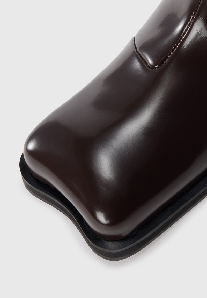 Bottine en cuir marron avec un bout arrondi, finition brillante et une semelle noire élégante. Détails de couture le long du haut.