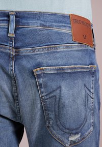 Denimové džíny s vybledlou modrou barvou, s oranžovým šitím a hnědou koženou nášivkou s nápisem "True Religion". Obsahují podrobné zadní kapsy.