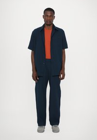Kurzärmliges, dunkelblaues Hemd mit Knöpfen über einem orangefarbenen T-Shirt, dazu passende dunkelblaue Hose und graue Turnschuhe. Einfaches Design, leichtes Material.