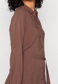 Brun, långärmad skjortklänning med knäppning fram, figursydd midja och krage. Tyget verkar slätt och lätt.