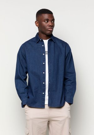 Mann trägt ein dunkelblaues Hemd mit Knöpfen über einem weißen T-Shirt und beigen Hosen, steht mit den Händen in den Taschen und schaut nach rechts.