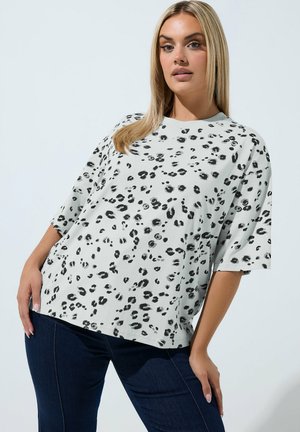 Blondynka w luźnej białej koszulce w czarne cętki lamparta i ciemnoniebieskich jeansach, pozująca na jednolitym jasnym tle.
