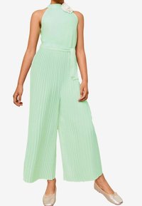 Miętowozielony kombinezon z plisowanym dołem, z halter neck i dopasowanym paskiem w talii; posiada biały akcent w postaci kwiatka z materiału. Jedwabista tekstura.