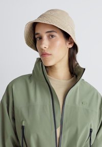 Grønn vanntett jakke med svarte glidelåser, kombinert med en beige bøttehatt. Jakken har høy krage og en glatt tekstur.