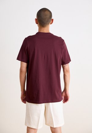 Mężczyzna z ogoloną głową, ubrany w jednolity bordowy T-shirt i białe szorty, stoi twarzą do białej ściany, z tatuażem na lewej ręce.