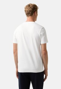 Blonde man in een eenvoudig wit T-shirt met korte mouwen en donkere broek, gezien vanaf de achterkant tegen een witte achtergrond.