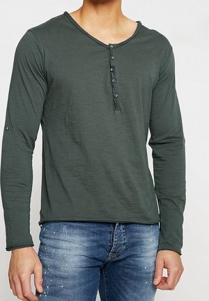 Homme portant un t-shirt henley à manches longues vert foncé avec des boutons, associé à un jean bleu délavé.