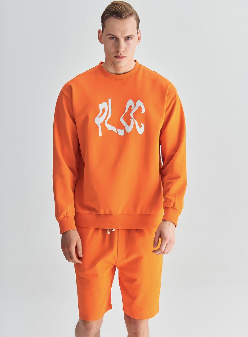 Pomarańczowy zestaw dresowy z bluzą i szortami, z wyraźnym białym logo, wykonany z gładkiego materiału, o luźnym kroju i ściągaczach na rękawach.