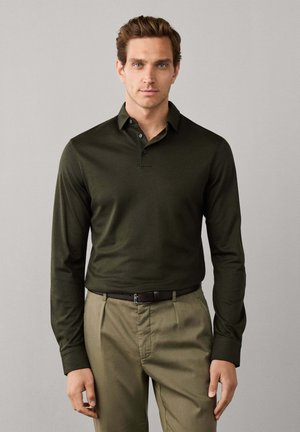 Man draagt een donkergroene langarmige polo en lichtbruine broek met een zwarte riem, staande tegen een effen lichte achtergrond.