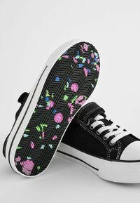 Sorte kanvas sneakers har hvide gummitåhætte og snørebånd. Sålen har et sort fiskebensmønster med farverige, abstrakte prikker.
