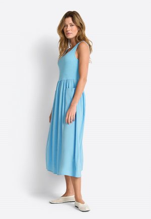 Frau steht seitlich, trägt ein ärmelloses hellblaues Midikleid mit Taschen und weiße gewebte Slip-On-Schuhe vor hellem Hintergrund.