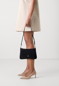 Borsa a tracolla nera in pelle con un emblema d'insetto dorato, forma slanciata e tracolla staccabile, tenuta da una persona che indossa un vestito beige a quadri.