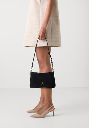 Borsa a tracolla nera in pelle con un emblema d'insetto dorato, forma slanciata e tracolla staccabile, tenuta da una persona che indossa un vestito beige a quadri.