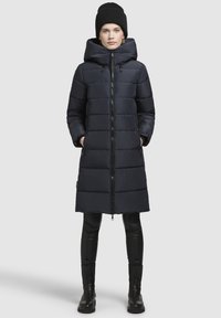 Navy puffer coat with a high collar, front zipper, and horizontal seams. Black leggings and boots complete the outfit. Beanie adds extra warmth.