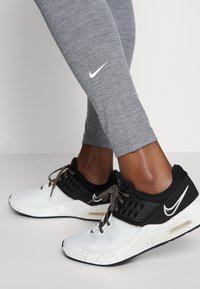 Nike sportovní boty s bílým síťovaným svrškem, černými akcenty a hnědou podrážkou s tečkami. Šedé legíny mají na lýtku bílou logo Nike.