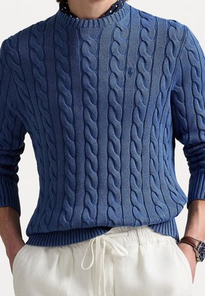 Mann trägt einen blauen Zopfpullover über einem gepunkteten Hemd, weiße Hosen mit Kordelzug und eine Armbanduhr.