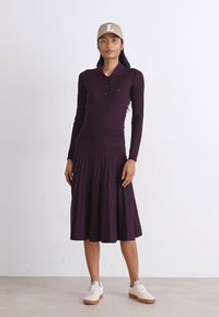 Ljubičasta rebrasta haljina s dugim rukavima, polo ovratnikom, tri dugmeta, suknja s nabranim detaljima, uparen s bijelim tenisicama i bež kapom.