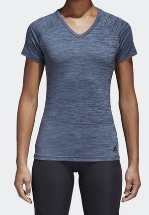 Femme portant un t-shirt de sport bleu ajusté à manches courtes et encolure en V, ainsi que des leggings noirs, debout les bras détendus sur les côtés.