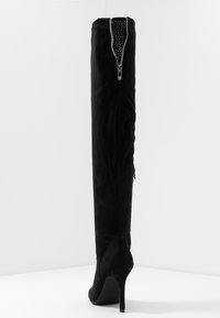 Bota de ante negro por encima de la rodilla con puntera puntiaguda, tacón de aguja alto y cierre decorativo con cremallera adornada con cristales en la parte trasera.