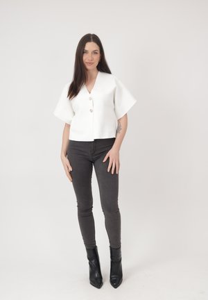 Femme aux longs cheveux foncés portant une blouse blanche boutonnée, un jean slim noir et des bottines noires sur un fond clair uni.