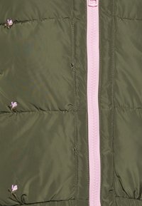 Tissu matelassé vert olive avec de petites fleurs brodées roses et une fermeture éclair rose centrale running verticalement.