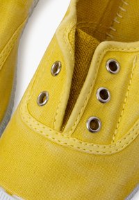 Zapatilla de lona amarilla con ojales de plata redondos, interior texturizado y costuras en contraste. Diseño ligero con una suela flexible.