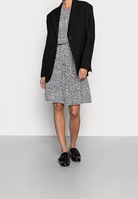 Blazer noir oversize superposé sur une robe à motif pied-de-poule blanc et noir, associé à des chaussures noires à enfiler. Textures lisses et motifs contrastés.