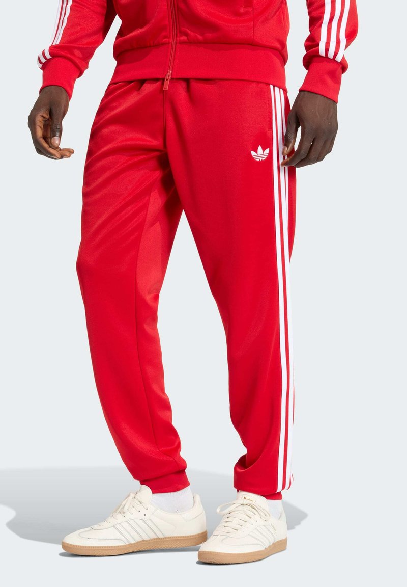 Άνδρας που φοράει κόκκινο παντελόνι φόρμας Adidas και ταιριαστό μπουφάν με λευκές ρίγες, σε συνδυασμό με λευκά αθλητικά παπούτσια και κάλτσες Adidas.