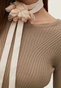 Eine Frau trägt ein beige geripptes Strickoberteil und ein cremefarbenes Stoffblumenaccessoire, das mit langen Bändern um ihren Hals gebunden ist.
