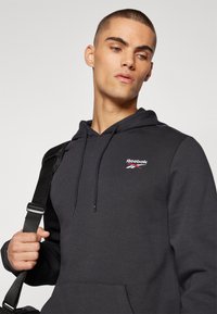 Sweat à capuche noir avec le logo Reebok. Comprend des cordons de serrage, une poche kangourou et une texture lisse. Modèle tenant un sac.