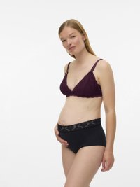 Έγκυος γυναίκα που φοράει μωβ δαντελένιο σουτιέν εγκυμοσύνης και μαύρα εσώρουχα με δαντέλα, κρατώντας τρυφερά την κοιλιά της, σε λευκό φόντο.