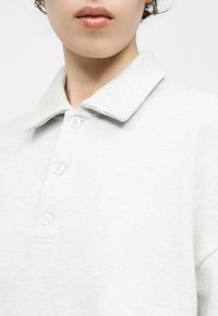 Osoba nosi svetlosivú polo košeľu s golierom a jemne vyšitým logom "Carhartt" na hrudi.