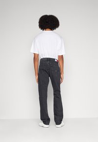Jeans de mezclilla negra con corte recto, que cuentan con bolsillos traseros y una etiqueta de logo. Combinados con una camiseta blanca básica y zapatillas blancas.