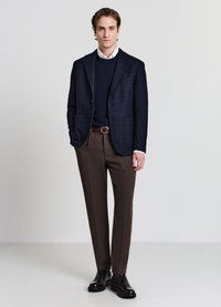 Blazer blu tessuto, maglione blu, camicia bianca, pantaloni marroni a fantasia, cintura marrone e stivali neri lucidi. In piedi su uno sfondo chiaro.