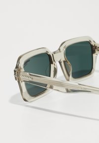 Ochelari de soare Carrera cu un cadru transparent de culoare bej deschis și lentile verzi. Formă pătrată cu brațe texturate ce prezintă detalii de branding.