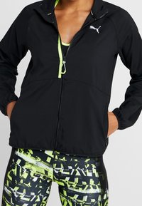 Henkilö, jolla on yllään musta Puma-takki, jossa on neonvihreää yksityiskohtaa, ja mustat sekä neonkuviolliset legginsit, kädet takin taskuissa.