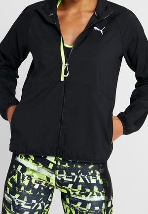 Persoon gekleed in een zwarte Puma-jas met neon groene accenten, over zwarte en neon patroonleggings, met handen in de zakken van de jas.