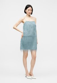 Νεαρή γυναίκα με κοντά μαύρα μαλλιά που φοράει μια αμάνικη μίνι φόρεμα ανοιχτού γαλάζιου χρώματος με φτέρες και λευκά τακούνια, στέκεται με το χέρι στη μέση.