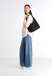 Sort tekstil skuldertaske med frontlomme, båret af en person iført en stribet skjorte, brede jeans og hvide sko.