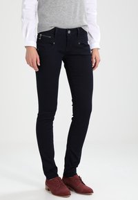 Vaqueros skinny azul oscuro de mezclilla, con un bolsillo con cremallera frontal y un detalle metálico. Combinados con un chaleco gris sobre una camisa blanca.