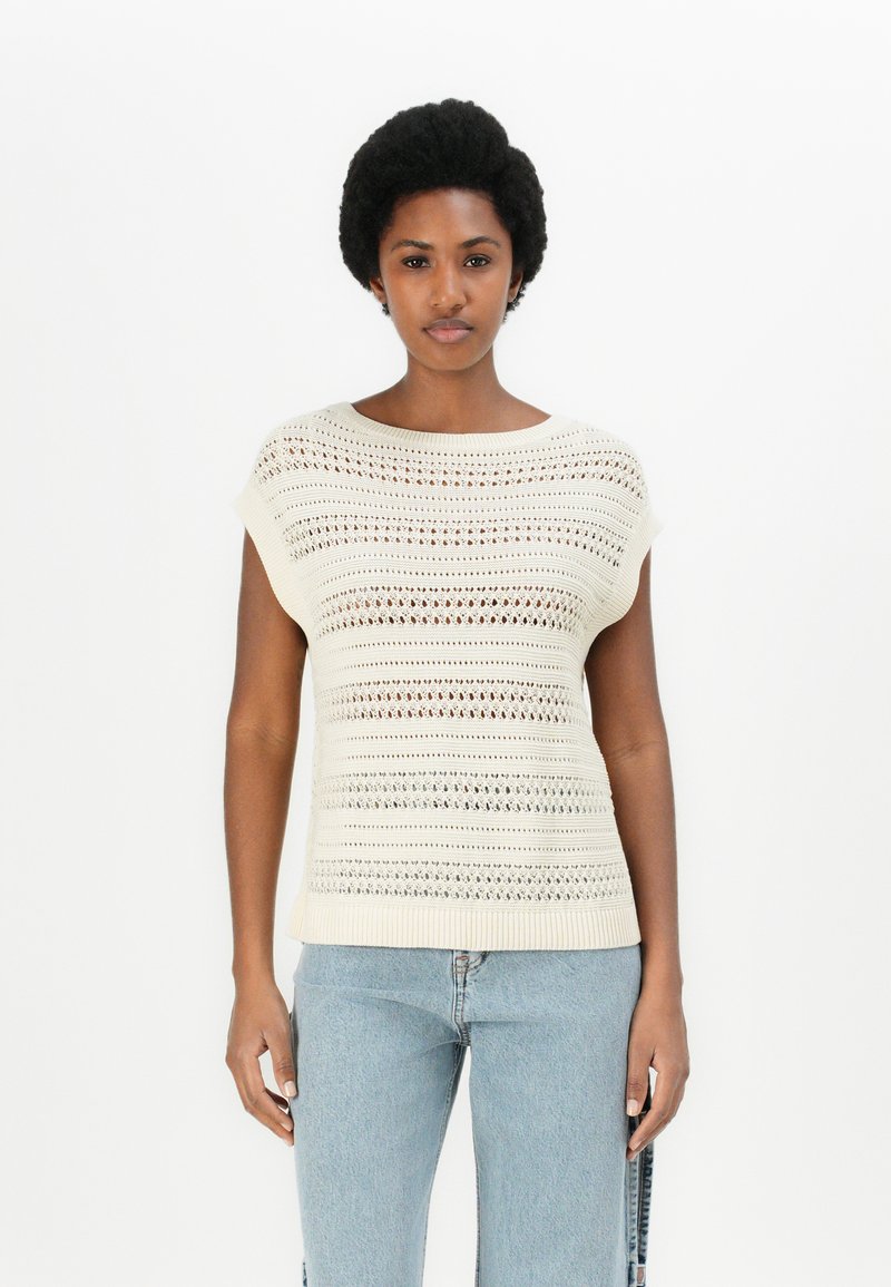 Lys beige strikket topp med korte ermer, med et teksturert åpent strikkemønster og ribbestrikket kant. Kombinert med lyseblå denimjeans.