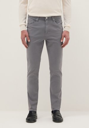 Man draagt grijze slim-fit broek, wit overhemd met lange mouwen en zwarte loafers, staand tegen een effen lichte achtergrond.