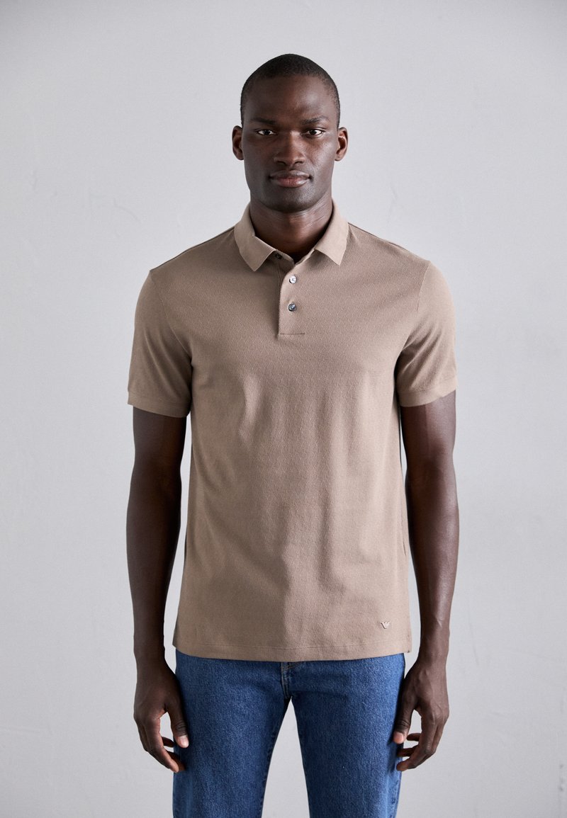 Chemise polo beige avec un motif texturé, patte de boutonnage à trois boutons et manches courtes, associée à un jean en denim bleu. Design et couleur simples.
