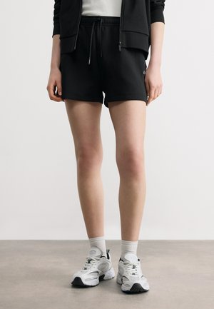 Osoba oblečená v čiernych šortkách so sťahovacou šnúrkou, čiernej vetrovke na zips, bielych ponožkách a bielych športových teniskách stojaca na sivom podlaží.