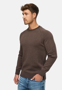 Un pull en maille marron avec un col rond et des poignets côtelés. Il présente une surface texturée, une coupe ajustée et est associé à un jean bleu clair.