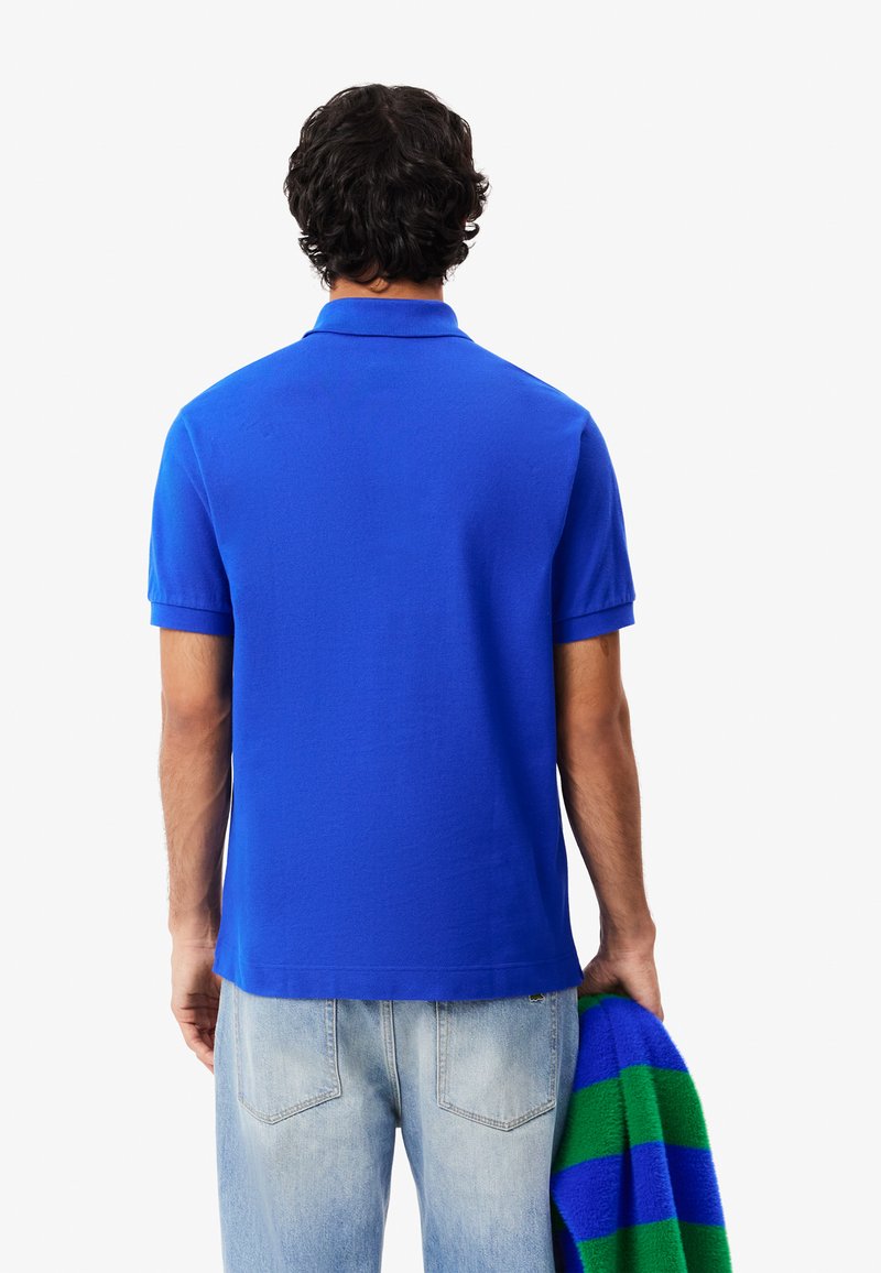 Blauses Polohemd mit kurzen Ärmeln, einem Kragen, einem geraden Saum, kombiniert mit hellblauen Jeans und hält einen grün gestreiften Pullover.