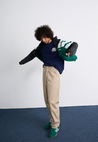 Men's sporty outfit featuring a black and green zip-up jacket, navy t-shirt, beige pants, and green sneakers with white stripes.